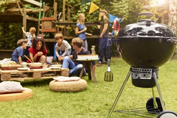 Ein Kugelgrill steht im Garten im Vordergrund. Im Hintergrund sitzen mehrere Personen an einem Tisch und unterhalten sich.